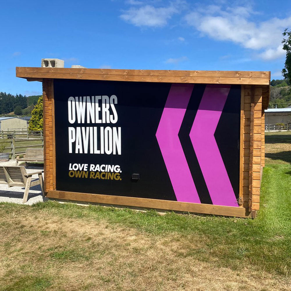 Wooden structure with 'Owners Pavilion' sign and racing-themed design outdoors.